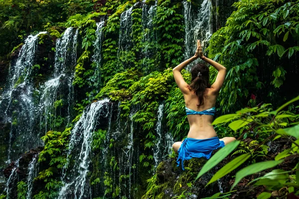 Женщина, сидящая на камне, практикующая йогу. Молодая женщина поднимает руки с мудрой намасте возле водопада. Banyu Wana Amertha водопад, Бали. Вид со спины . — стоковое фото