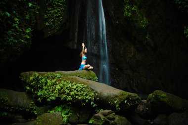 Genç beyaz kadın meditasyon yapıyor, şelalede yoga yapıyor. Namaste Mudra 'da eller havaya. Leke Leke Şelalesi, Bali, Endonezya. 