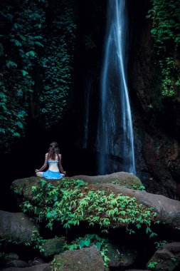 Genç bir kadın kayanın üzerinde oturuyor, şelalenin yanında yoga yapıyor. Eller Gyan Mudra 'da. Leke Leke Şelalesi, Bali. Arkadan görüntüle.