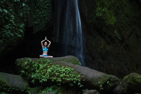 Young Caucasian woman meditating, practicing yoga at waterfall. Hands raising up in namaste mudra. Leke Leke waterfall, Bali, Indonesia.