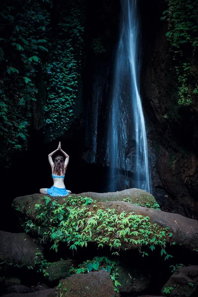 Donna seduta sulla roccia, che pratica yoga. Giovane donna che alza le braccia con namaste mudra vicino alla cascata. Cascata Leke Leke, Bali. Vista da dietro . — Foto Stock