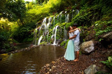 Mutlu çok ırklı çift sarılıp öpüşüyor. Tropiklerde şelalenin yanında aşık bir çift. Romantik bir ilişki. Wana Amertha Şelalesi Wanagiri, Bali
