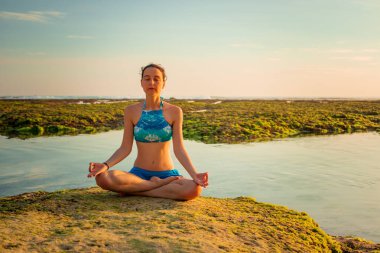 Genç bir kadın, meditasyon yapıyor, yoga yapıyor ve plajda pranayama yapıyor. Günbatımı yoga antrenmanı. Eller Gyan Mudra 'da. Melasti plajı, Bali