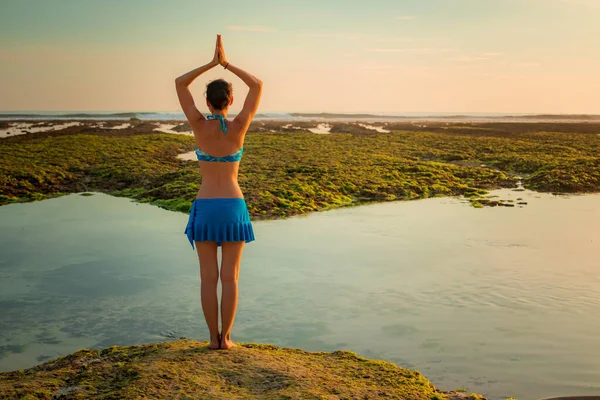 Yoga pose. Woman standing on the sand, practicing yoga. Young woman raising arms with namaste mudra at the beach. Bali. View from back.