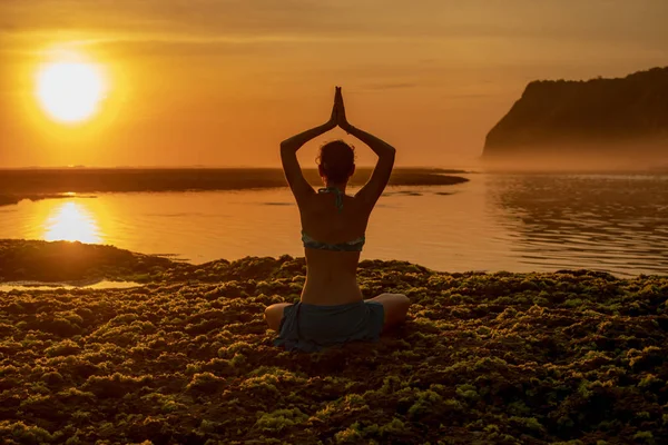 Genç bir kadın meditasyon yapıyor, yoga yapıyor ve plajda pranayama yapıyor. Günbatımı yoga antrenmanı. Namaste Mudra 'da eller havaya. Arkadan bak. Melasti plajı, Bali — Stok fotoğraf