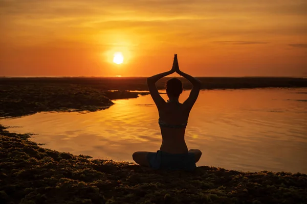 Yoga pozu. Kumsalda oturan bir kadın, yoga yapıyor. Gün batımının altın saatinde namaste Mudra ile kollarını kaldıran genç kadın. Arkadan bak. Melasti plajı, Bali. 