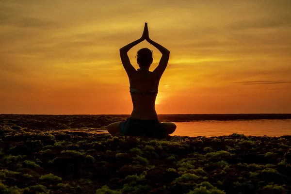 Yoga pose. Woman sitting on the beach, practicing yoga. Young woman raising arms with namaste mudra during sunset golden hour. View from back. Melasti beach, Bali.