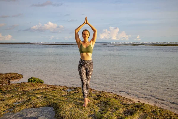 Yoga pose. Woman practicing yoga at the beach. Young woman standing and raising arms with namaste mudra. Healthy lifestyle. Yoga retreat in Bali, Melasti beach.