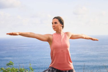 Savaşçı pozunu yakın çekim. Yoga yapan genç bir kadın, Virabhadrasana pozunda duruyor. Uçurumda açık hava yogası. Çalış. Sağlıklı bir yaşam tarzı. Arka planda okyanus. Yoga geri çekilme, Bali.