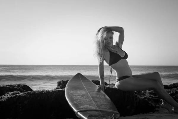 Monochroom. Surfer meisje zit op de rots. Blauw longboard. Surfen levensstijl. Bali, Indonesië — Stockfoto