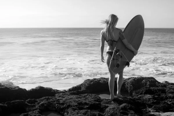 Monokrom. Surfare flicka med surfbräda gå på stranden. Ung sexig kvinna klädd i svart bikini. Utsikt från bak. Bali, Indonesien — Stockfoto