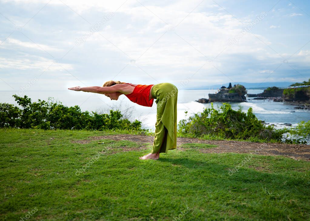 Young woman practicing yoga, standing in Ardha Uttanasana pose. Standing half forward bend. Back stretching. Outdoor yoga on the cliff. Yoga retreat. Tanah Lot temple Bali, Indonesia