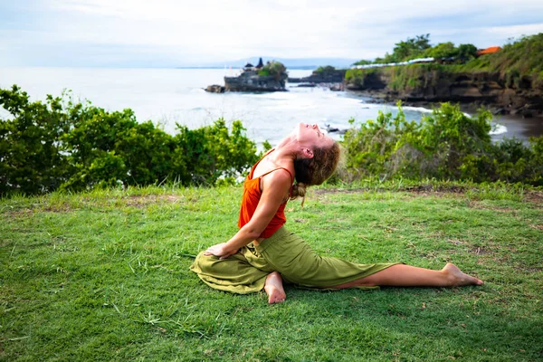 Outdoor yoga practice. Young woman practicing Ardha Kapotasana, Half Pigeon Pose. Chest opener improving breathing. Healthy lifestyle. Yoga retreat. Tanah Lot temple, Bali, Indonesia