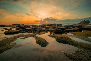 Seascape. Beach with rocks and stones. Low tide. Sun and clouds reflection in water. Slow shutter speed. Soft focus. Tanah Lot beach, Bali, Indonesia