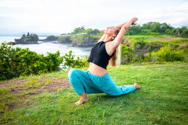 Outdoor yoga. Young woman practicing Ashwa Sanchalanasana, Equestrian or riding pose as part of the Sun Salutation series of asanas. Hands raising in namaste mudra. Yoga retreat. Tanah Lot temple Bali