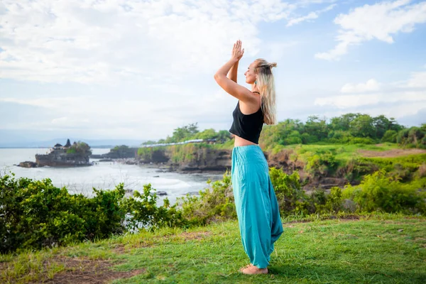 Outdoor yoga practice. Young woman standing on the grass. Hands raising  up in namaste mudra, closed eyes. Calm and peaceful mood. Yoga retreat. Cliff near Tanah Lot temple, Bali