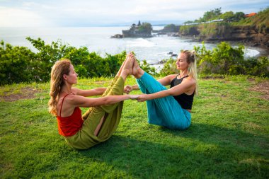 İki genç kadın Ubhaya Padangusthasana, Geniş Açı Ön Kıvrım antrenmanı yapıyor. Bacaklarını ve çekirdeğini güçlendir. Açık hava sağlığı. Sağlık sigortası konsepti. Yoga inzivası. Tanah Lot Tapınağı, Bali. 
