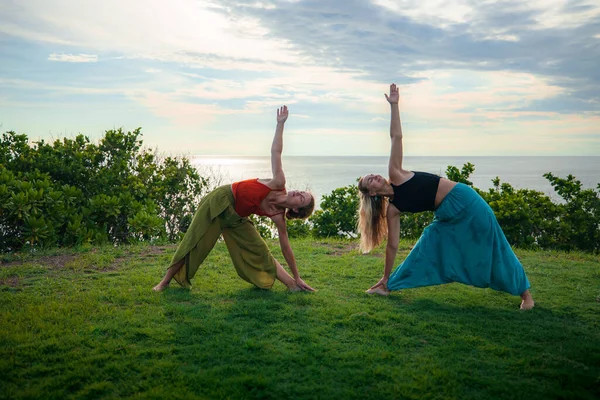 Outdoor yoga practice. Two young women practice Utthita Trikonasana, Extended Triangle Pose. Standing asana. Healthcare concept. Yoga retreat. Tanah Lot temple, Bali, Indonesia