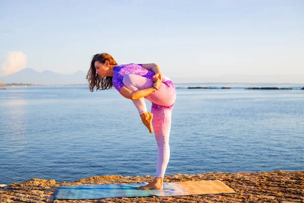 Çekici bir kadın, gün doğumunda yoga antrenmanına hazırlanıyor. Okyanus kenarında açık hava yoga antrenmanı. Sağlık ve kişisel bakım konsepti. Tek ayak üzerinde durarak dengeyi sağlayın. Bali, Endonezya