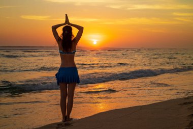 Yoga pozu. Sahilde durmuş yoga yapan bir kadın silueti. Gün batımının altın saatinde namaste Mudra ile kollarını kaldıran genç kadın. Arkadan bak. Güneş ışığı yansıması. Melasti plajı, Bali.