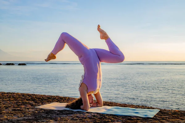 Okyanus kenarında gün doğumu yoga antrenmanı. Salamba Shirshasana, Yoga Headstand pratiği yapan genç bir kadın ters dönmüş bir asana. Kişisel bakım konsepti. Yoga inzivası. Bali, Endonezya
