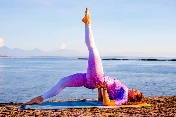 Outdoor yoga practice near the ocean. Attractive woman practicing variation of Setu Bandhasana, Shoulder supported bridge pose with one leg up. Healthy life. Yoga retreat. Bali, Indonesia