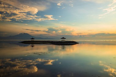 Gün doğumu deniz manzarası. Dağlarla ve Agung volkanıyla kaplı bir manzara. Okyanustaki yapay bir adada geleneksel çardaklar. Su yansıması. Sakin su yüzeyi. Yumuşak odaklanma. Sanur Plajı, Bali, Endonezya.