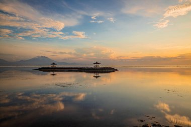Gün doğumu deniz manzarası. Dağlarla ve Agung volkanıyla kaplı bir manzara. Okyanustaki yapay bir adada geleneksel çardaklar. Su yansıması. Sakin su yüzeyi. Yumuşak odaklanma. Sanur Plajı, Bali, Endonezya.