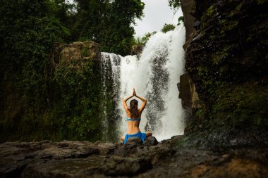 Genç beyaz kadın kayanın üzerinde oturuyor, meditasyon yapıyor, yoga yapıyor ve pranayama yapıyor. Eller namaste mudra 'da. Tegenungan şelalesi. Yoga inzivası. Kişisel bakım konsepti. Arkadan bak. Ubud, Bali