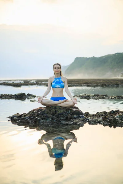 Young woman, meditating, practicing yoga and pranayama at the beach. Sunset yoga practice. Hands in gyan mudra. Water reflection. Healthy lifestyle. Yoga retreat. Melasti beach, Bali, Indonesia