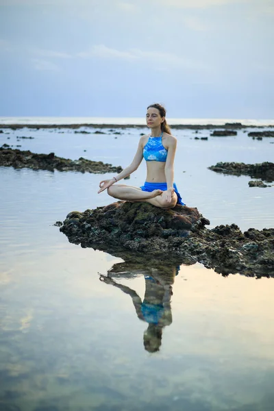 Young woman, meditating, practicing yoga and pranayama at the beach. Sunset yoga practice. Hands in gyan mudra. Water reflection. Support immune system. Yoga retreat. Melasti beach, Bali, Indonesia