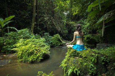 Genç beyaz kadın kayanın üzerinde oturmuş yoga ve pranayama yapıyor. Eller Gyan Mudra 'da. Arkadan bak. Asya 'da tatil. Sakin ve rahatlatıcı bir atmosfer. Tropik doğa manzarası. Bangli, Bali.