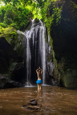 Genç beyaz kadın şelalenin önünde duruyor, meditasyon yapıyor, yoga yapıyor ve pranayama yapıyor. Namaste mudra. Arkadan bak. Sağlıklı bir yaşam tarzı. Krisik Şelalesi Bangli, Bali, Endonezya 'da.