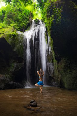 Vrikshasana asana, ağaç duruşu. Genç beyaz kadın meditasyon yapıyor, şelalede yoga yapıyor. Namaste Mudra 'da eller havaya. Arkadan bak. Krisik Şelalesi Bangli, Bali, Endonezya 'da.