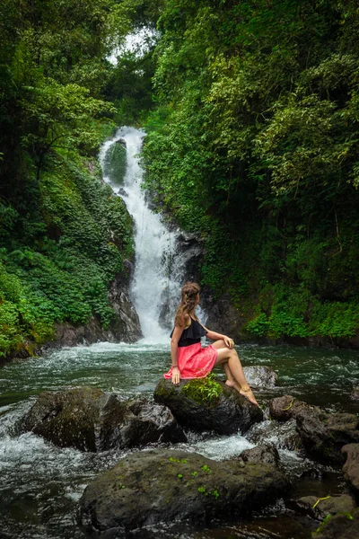 Genç beyaz kadın kayada oturuyor ve şelale manzarasının tadını çıkarıyor. Seyahat yaşam tarzı. Siyah ve pembe elbiseli bir kadın. Sambangan 'da Dedari Şelalesi, Bali, Endonezya