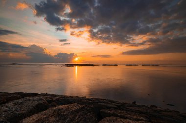 Gün doğumu deniz manzarası. İnanılmaz bir su yansıması. Bulutlu gökyüzü. Gün ışığı ufukta. Renkli gün doğumu. Taşlı ön plan. Sakin su yüzeyi. Nusa Dua Sahili, Bali, Endonezya.