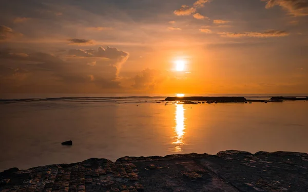 Rising sun at horizon. Sunrise seascape. Amazing water reflection. Cloudy sky. Bright sunlight at horizon. Foreground with stones. Nusa Dua beach, Bali, Indonesia.