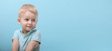 Mavi Polo giyen üç yaşındaki bir çocuk kameraya poz veriyor ve farklı duygular gösteriyor. Çocukların duyguları. Mavi arka plandaki çocuklar. Çocuk düşünür ve yüzüne parmağını tutar.