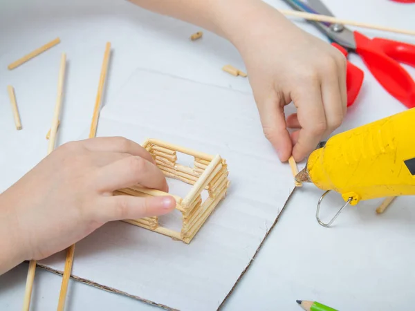 Bambu çubuklarından ev yapmak için adım adım talimatlar. Çocukların yaratıcılığı, el işi, online dersler. Beşinci adım tutkal tabancasıyla, çocuk bambu çubukları takıyor.