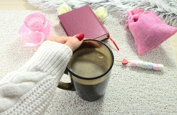 Female hand with a cup in hand, a book on the table. Relax on a weekend with a mug of espresso.Romantic photo with pink candles.