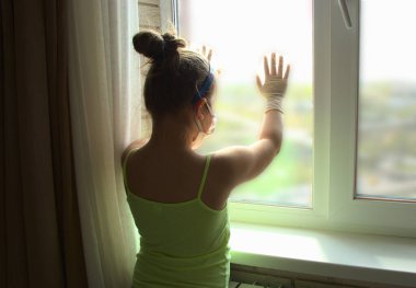 Eldivenli ve maskeli bir kız pencereden dışarı bakıyor. Çocuk pencerenin yanında üzgün, karantina altında..