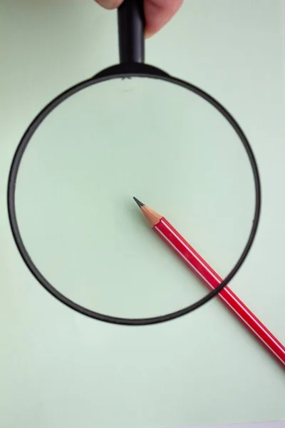 Una matita rossa sotto una lente d'ingrandimento primo piano su uno sfondo chiaro . — Foto Stock