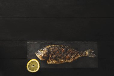 Siyah ahşap arka planda bir dilim limonla siyah bir taşın üzerinde taze balık ya da balık pişirilmiş.