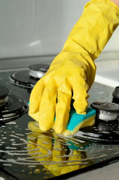 A hand in a yellow rubber glove washes a gas stove on a sunny day. Kitchen cleaning