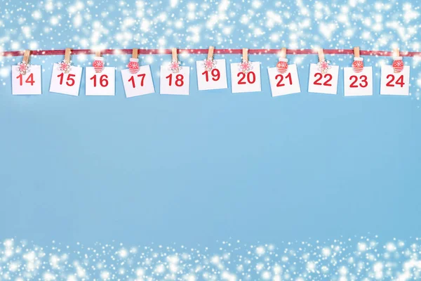 Advent takviminin 14-24 bölümü. Şarjörlerde numaralar olan çarşaflar mavi arkaplanda kırmızı kurdelede asılı..