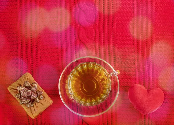 Valentinstag heimelige gemütliche Kulisse. Tasse Tee, Plüschherz, Geschenkbox auf rotem Strickpullover mit Bokeh. Ansicht von oben. — Stockfoto