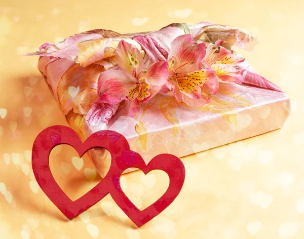 Trendy verpackt in Furoshiki-Technik Geschenkbox mit Blumen Alstroemeria und zwei rote Holzherzen auf gelb mit Bokeh. — Stockfoto