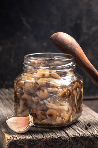 Glass jar with homemade pickled forest mushrooms honey agarics, wooden spoon and garlic on old wooden board on dark backdrop. Fermented food. Sustainable food. Close-up, vertical orientation.