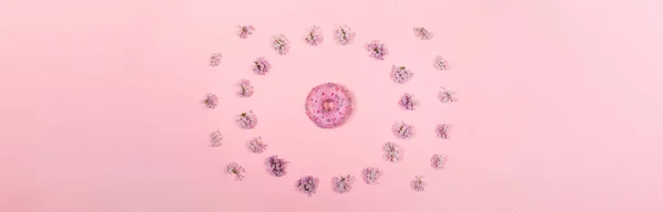 Creative banner with one traditional donghnut with lilac glaze surrounded with flowers of lilac on pink background . — стоковое фото