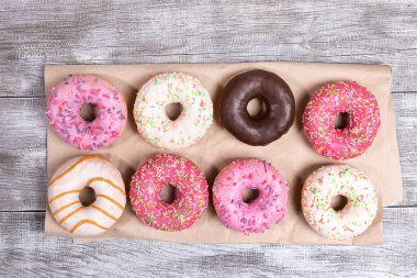 Eight traditional doughnuts with multicolored glaze neatly laid out on wrapping paper on white painted wooden background. Donuts - traditional sweet pastry. Top view, flat lay. clipart